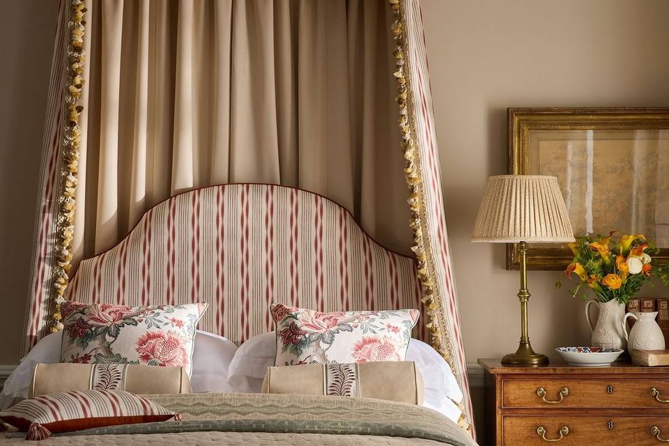 A striped canopy headboard with a bedside lamp and a jug of flowers in a country bedroom