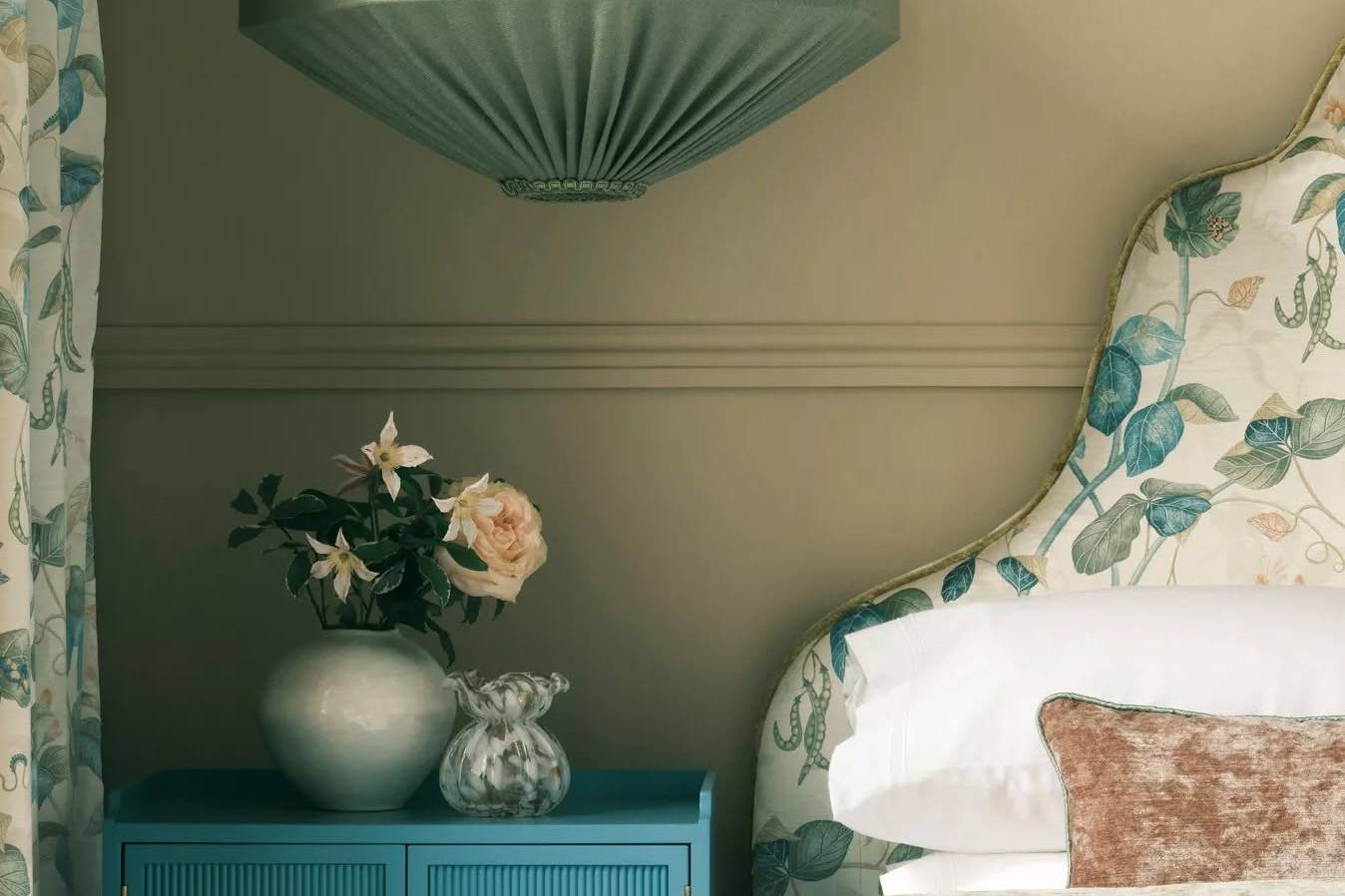 A floral upholstered chair beside a vase of roses in a green painted sitting room with patterned curtains